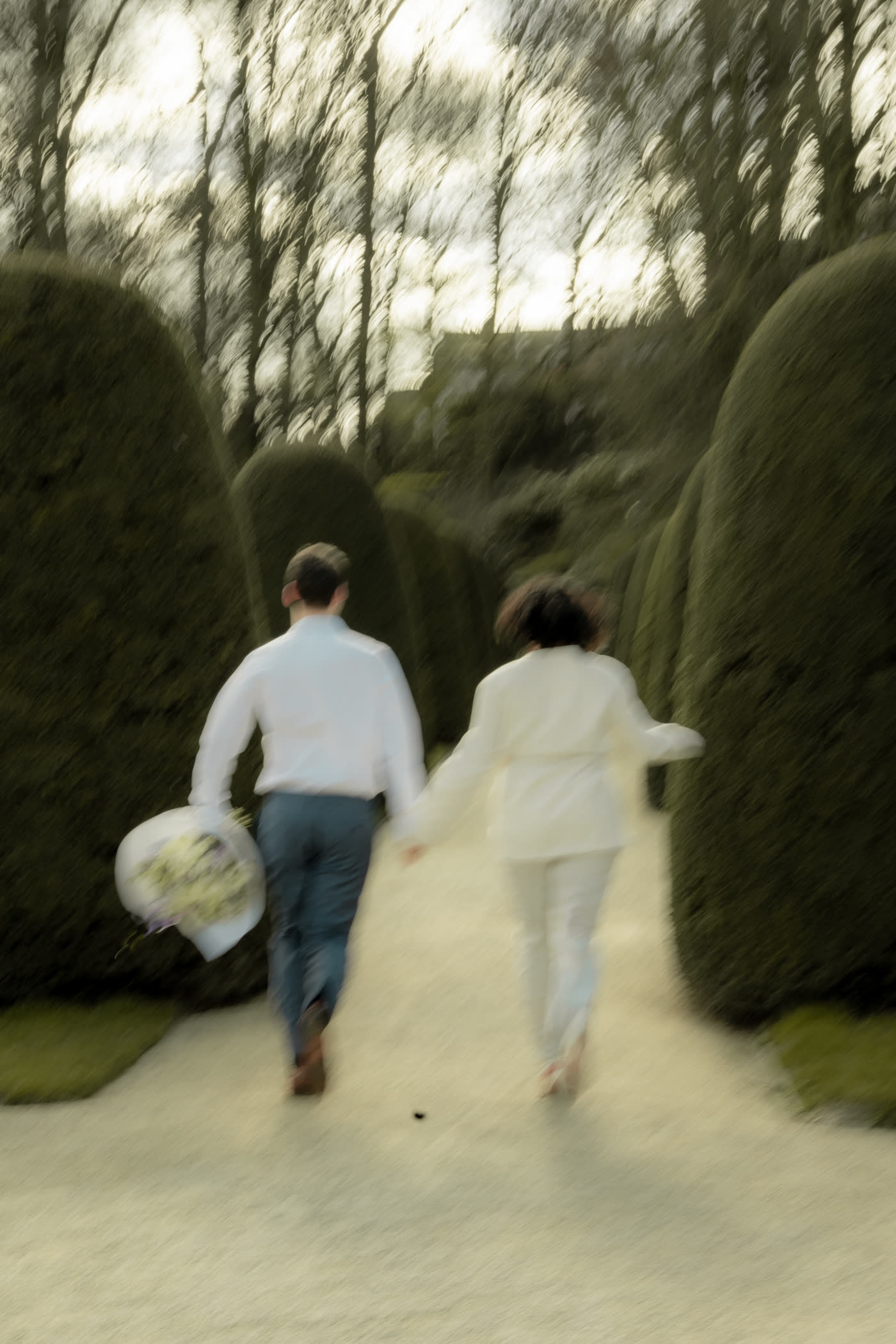 Simon et Laura de dos, courant main dans la main sur une allée bordée de haies, un bouquet à la main